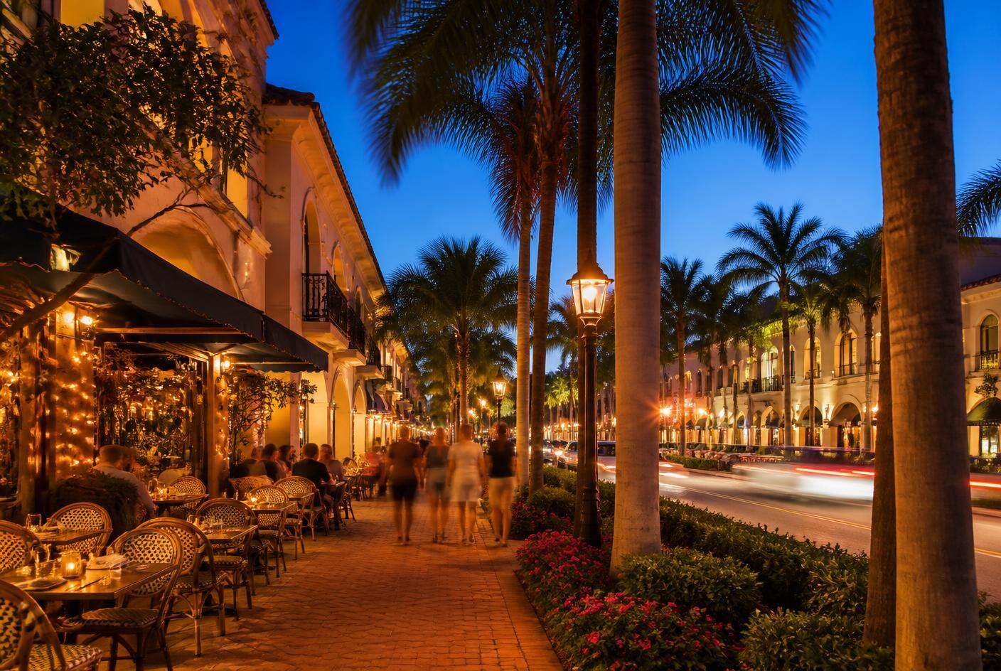 Palm-lined Naples street at dusk with lit outdoor cafe tables and brick sidewalks